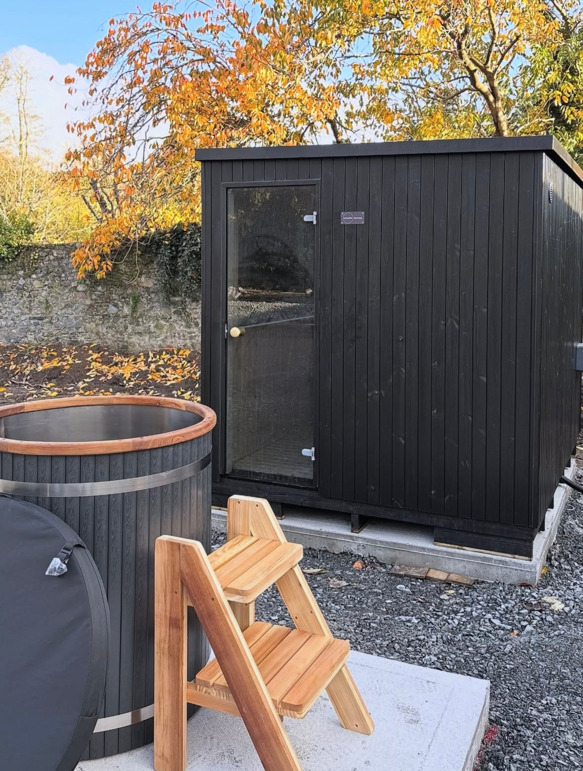 Black wooden shed with a hot tub and wooden steps in an outdoor setting.