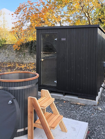 Black wooden shed with a hot tub and wooden steps in an outdoor setting.