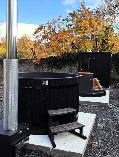 Black hot tub with steps outdoors against a backdrop of trees with autumn foliage.